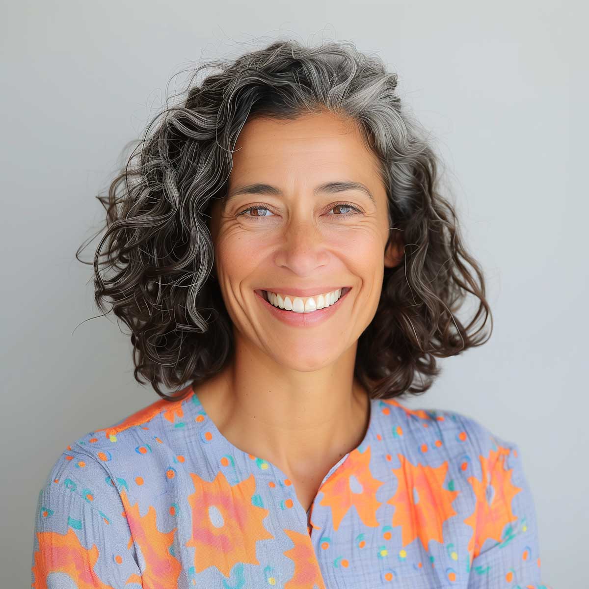 Lächelnde Frau mit lockigem, grauem Haar und einem bunten Oberteil vor einem hellen Hintergrund.