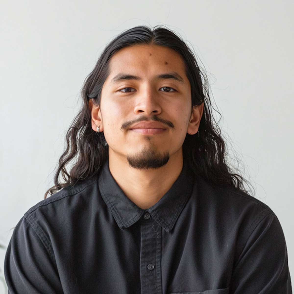 Junger Mann mit langen, lockigen Haaren und leichtem Bart trägt ein schwarzes Hemd und schaut direkt in die Kamera.