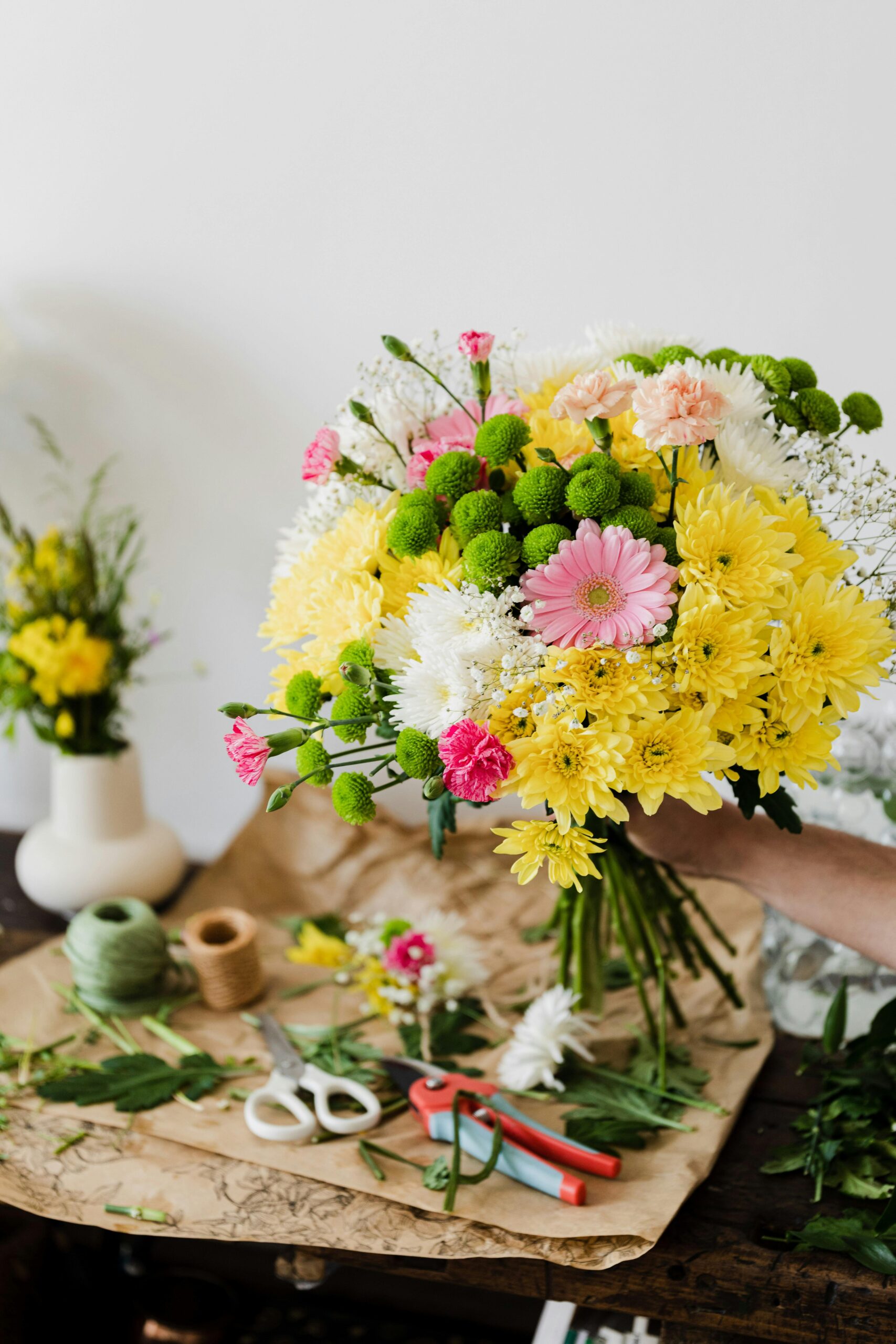 Ein bunter Blumenstrauß mit gelben, pinken und weißen Blumen wird in einer Hand gehalten, während im Hintergrund Werkzeug und weitere Blumen zur Gestaltung bereitliegen.